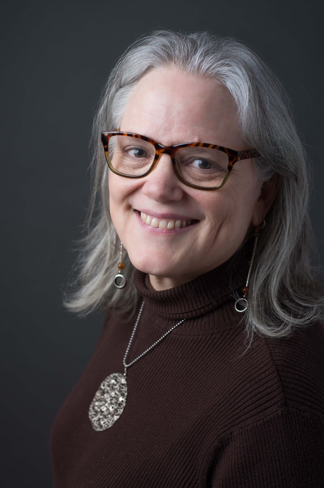 Dr. Janice Campbell in a brown, ribbed, turtleneck, shoulder length grey hair, tortoise shell framed glasses, a large silver pendant necklace and long silver earrings. She is smiling and looking at the camera over her left shoulder.